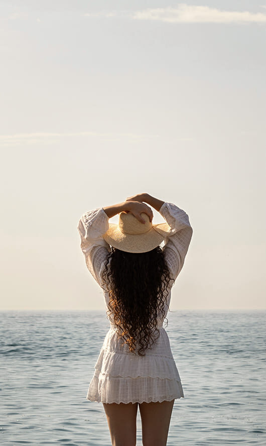 Woman with long curly hair and straw hat gazing at the sea at Mikri Santa Maria beach.